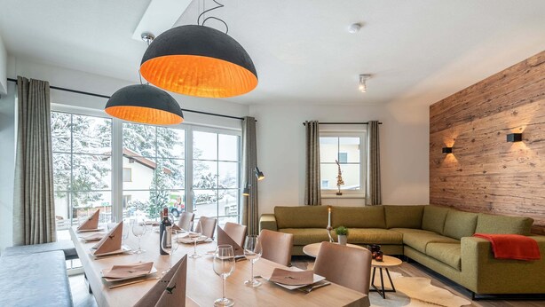 A modern dining area with a long wooden table, multiple chairs, and pendant lighting. There is a green sofa and a small round table with a plant and candle in the living area. - Luxury Chalet with Sauna near Ski Area in Salzburg (Sankt Johann im Pongau)