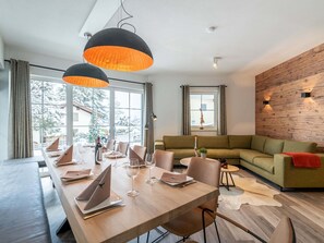 A modern dining area with a long wooden table, multiple chairs, and pendant lighting. There is a green sofa and a small round table with a plant and candle in the living area. - Luxury Chalet with Sauna near Ski Area in Salzburg (Sankt Johann im Pongau)