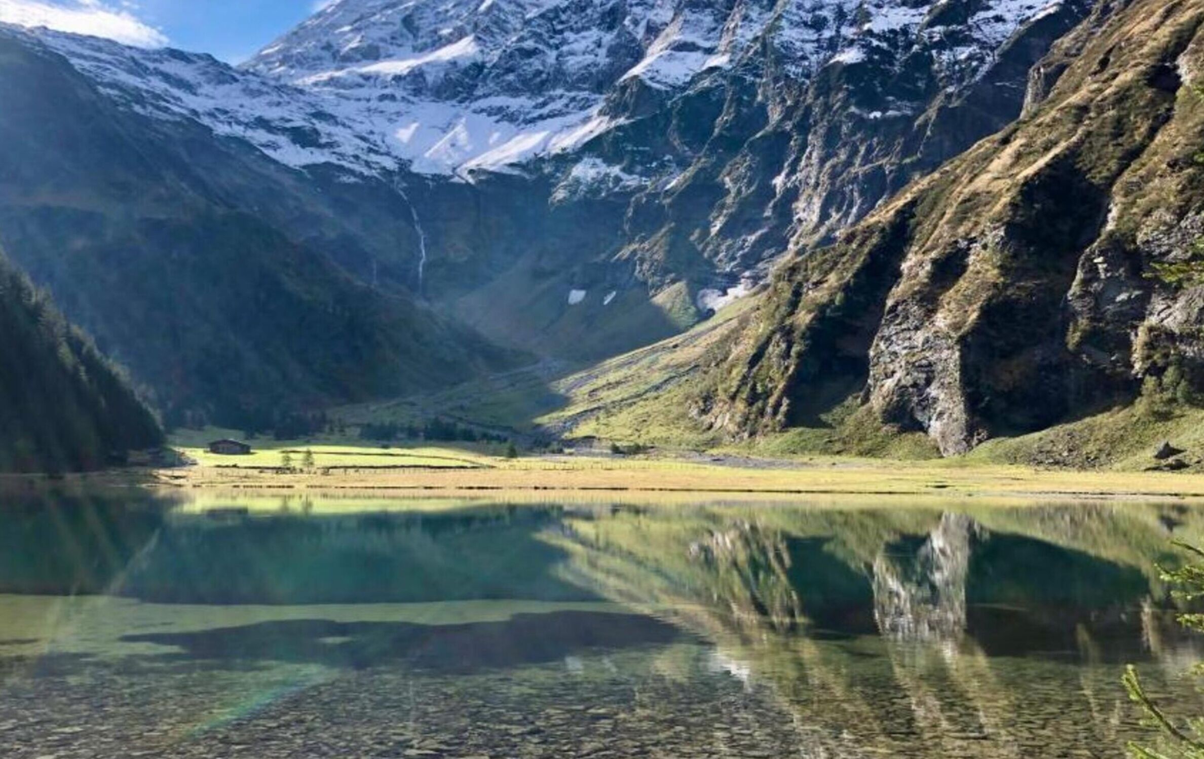 A clear mountain lake with reflections, surrounded by rugged mountains and greenery.