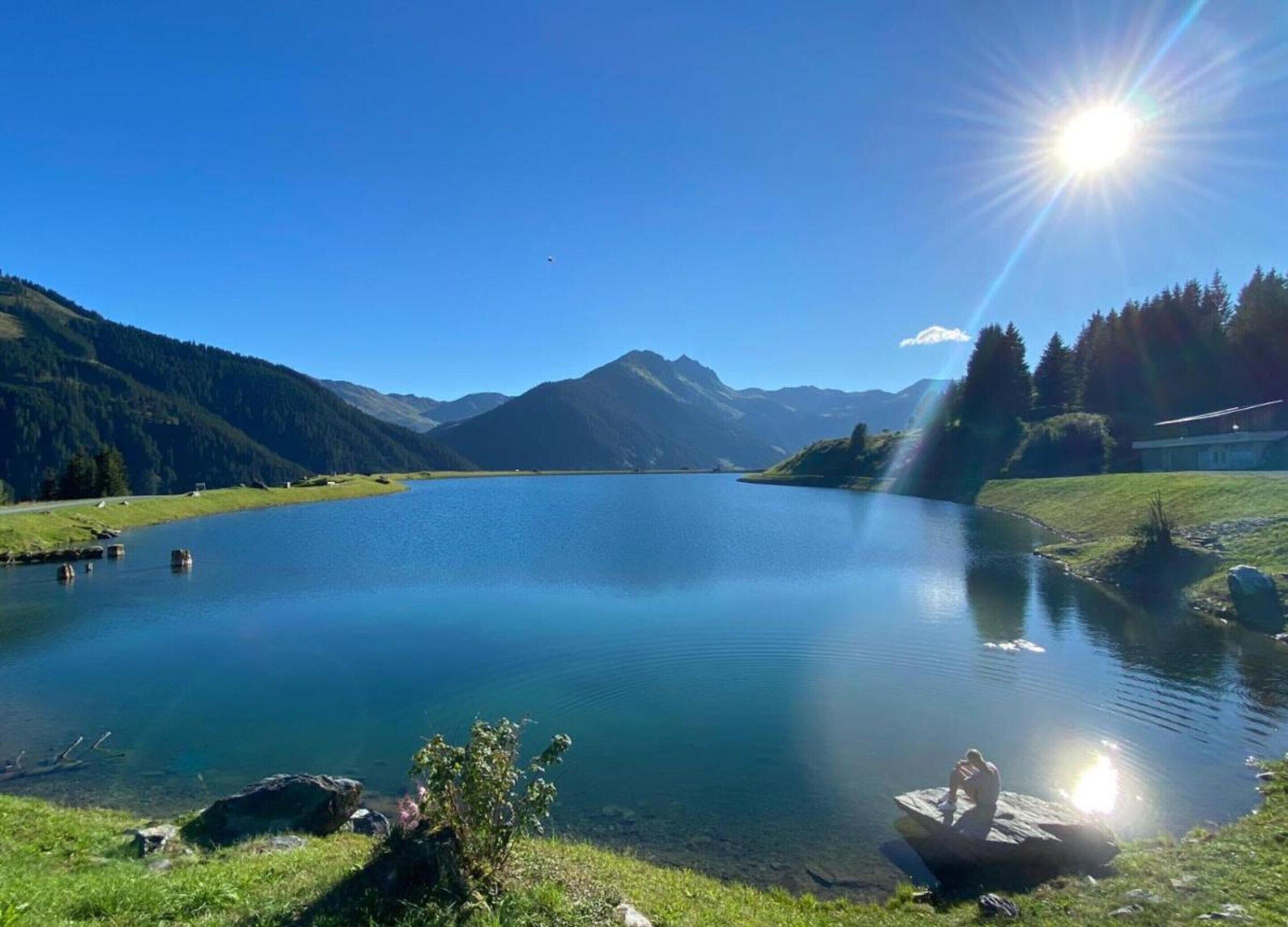 Un lago sereno rodeado de montañas, con cielo despejado y un sol brillante.