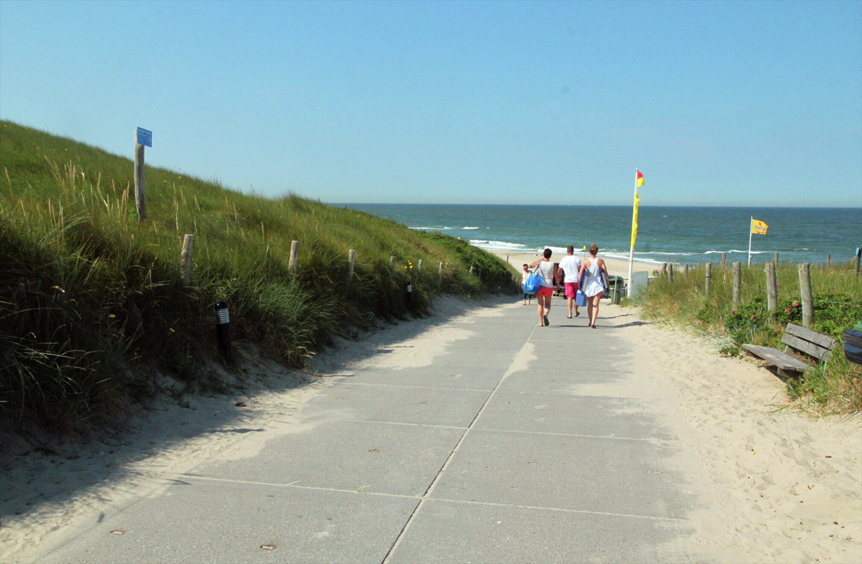 A paved pathway beside a beach with grassy dunes and a clear sky.