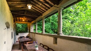 Un porche techado al aire libre con mesas y sillas de madera, un jarrón con flores y vistas a follaje exuberante.