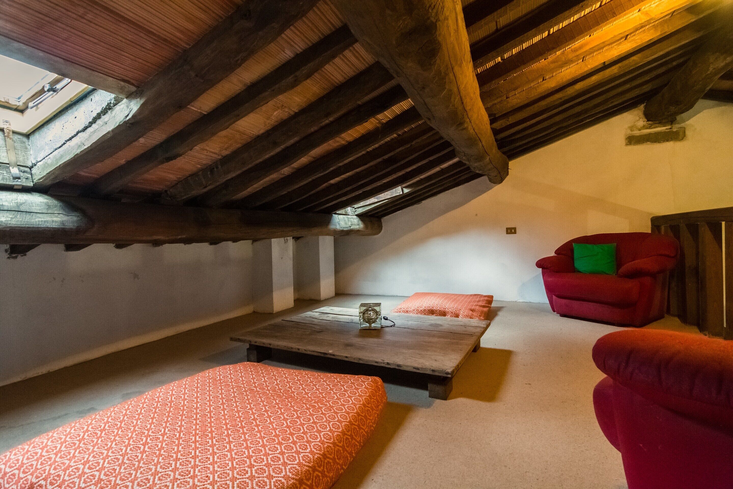 Une pièce mansardée chaleureuse avec un plafond en bois, une petite table avec une lampe, deux lits à motifs et un fauteuil rouge.