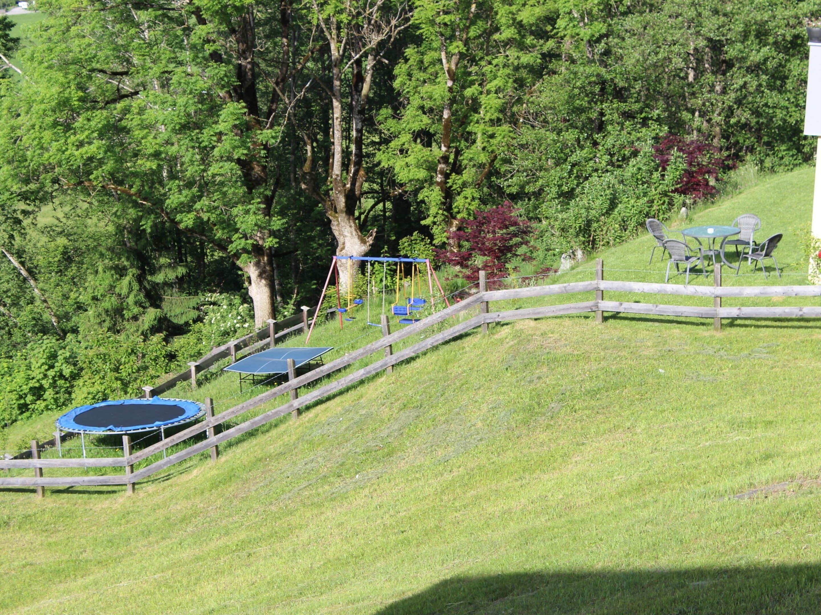 Une colline herbeuse, avec une clôture en bois, un trampoline et une balançoire.