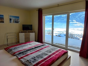 A bedroom with a bed, a TV on a dresser, a window with a snowy mountain view, and a red curtain. - Sunlit Apartment near Ski Area in Walchen (Piesendorf)