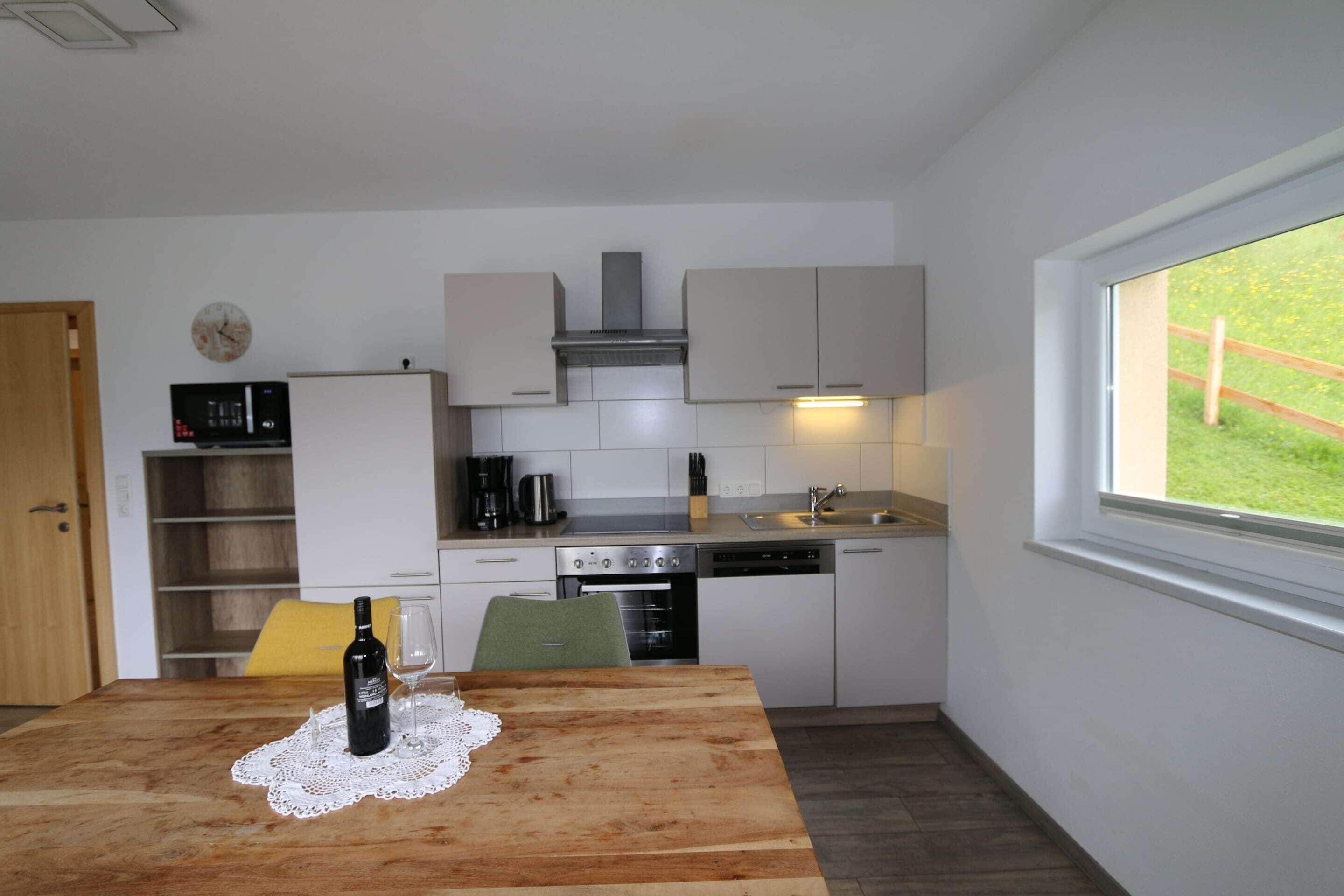 Una cucina compatta con mobili bianchi, un forno a microonde, un fornello e una lavastoviglie. C'è un tavolo di legno con una bottiglia e un bicchiere.