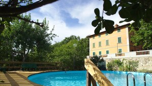 A wooden deck with a pool, surrounded by trees and a building with green shutters. - Hillside Villa in the Heart of Chianti (San Casciano in Val di Pesa)
