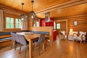 A log cabin interior with a wooden dining table, gray chairs, a bench, a red kitchen area, and a fireplace.