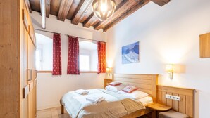 Un dormitorio con una cama de madera, una mesita de noche, un banco, una ventana con cortinas rojas y una lámpara fijada a la pared.