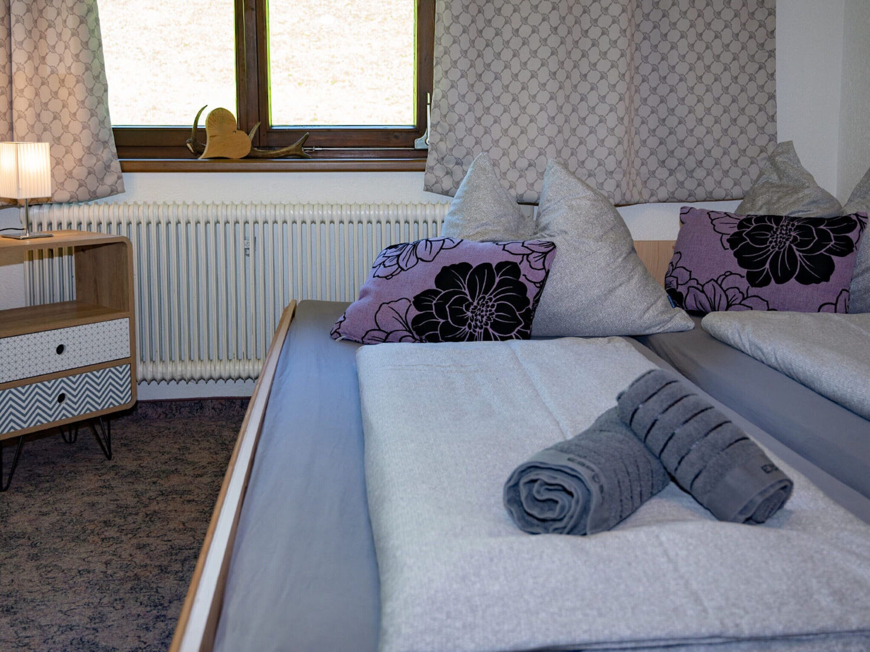 A neatly made bed with rolled towels, a bedside table with a lamp, and patterned curtains.