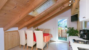 Une cuisine chaleureuse avec un coin repas, des poutres en bois au plafond, un sol carrelé et une vue sur un jardin, visible à travers une porte vitrée.