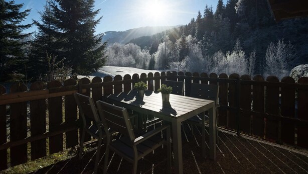 Een houten terras met een tafel en stoelen, met uitzicht op een bebost dal met een heldere lucht en de zon die schijnt.