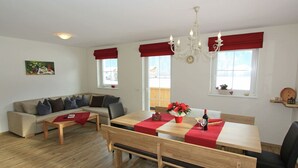 A modern living room with a sofa, a dining table with benches, a chandelier, and large windows with red curtains. - Apartment in Aschau Near Hochzillertal Slopes (Aschau Im Zillertal)