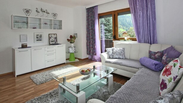 Una sala de estar moderna con una mesa de centro de vidrio, sofá blanco y vistas al exterior a través de una ventana con cortinas moradas.