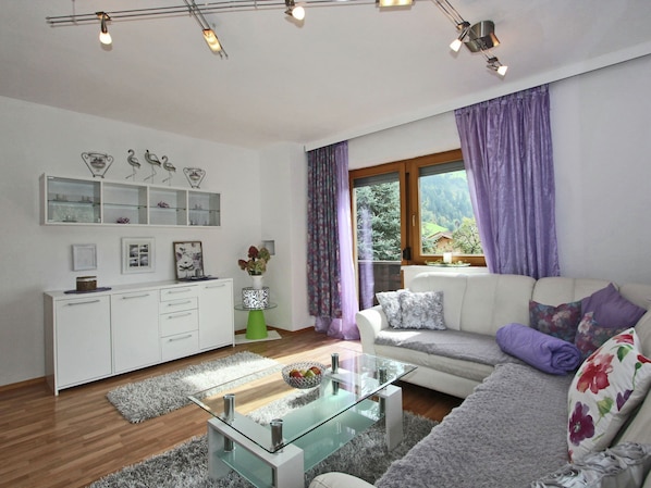 Un salon moderne avec une table basse en verre, un canapé blanc et une vue sur l’extérieur grâce à une fenêtre ornée de rideaux violets.