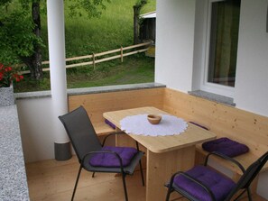 Un petit espace extérieur aménagé avec une table et des chaises en bois, une nappe en dentelle blanche, et une vue sur une pelouse et une clôture en bois.