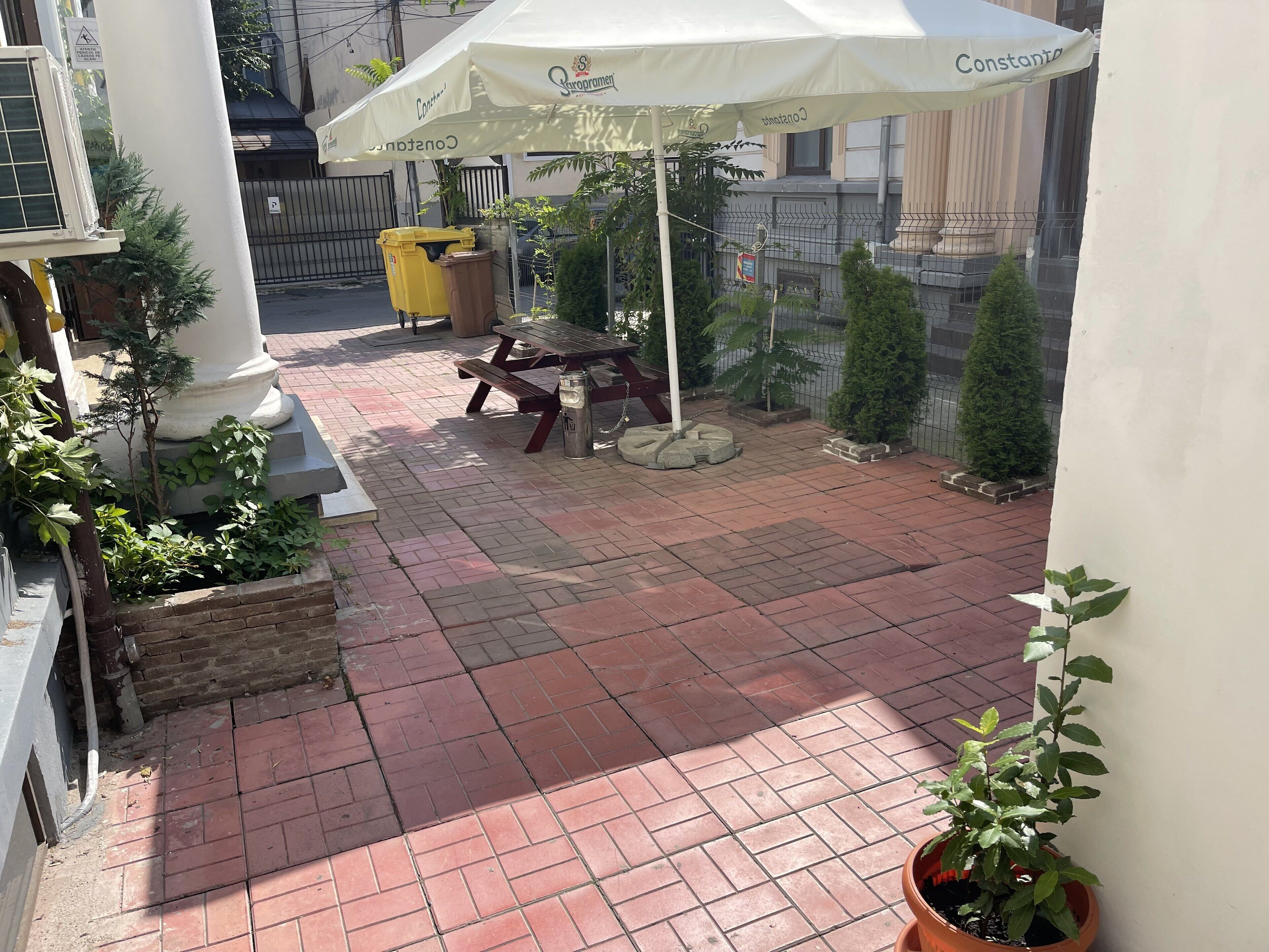 Eine Terrassenfläche mit Picknicktisch und Mülleimer, umgeben von Topfpflanzen und einem weißen Sonnenschirm.
