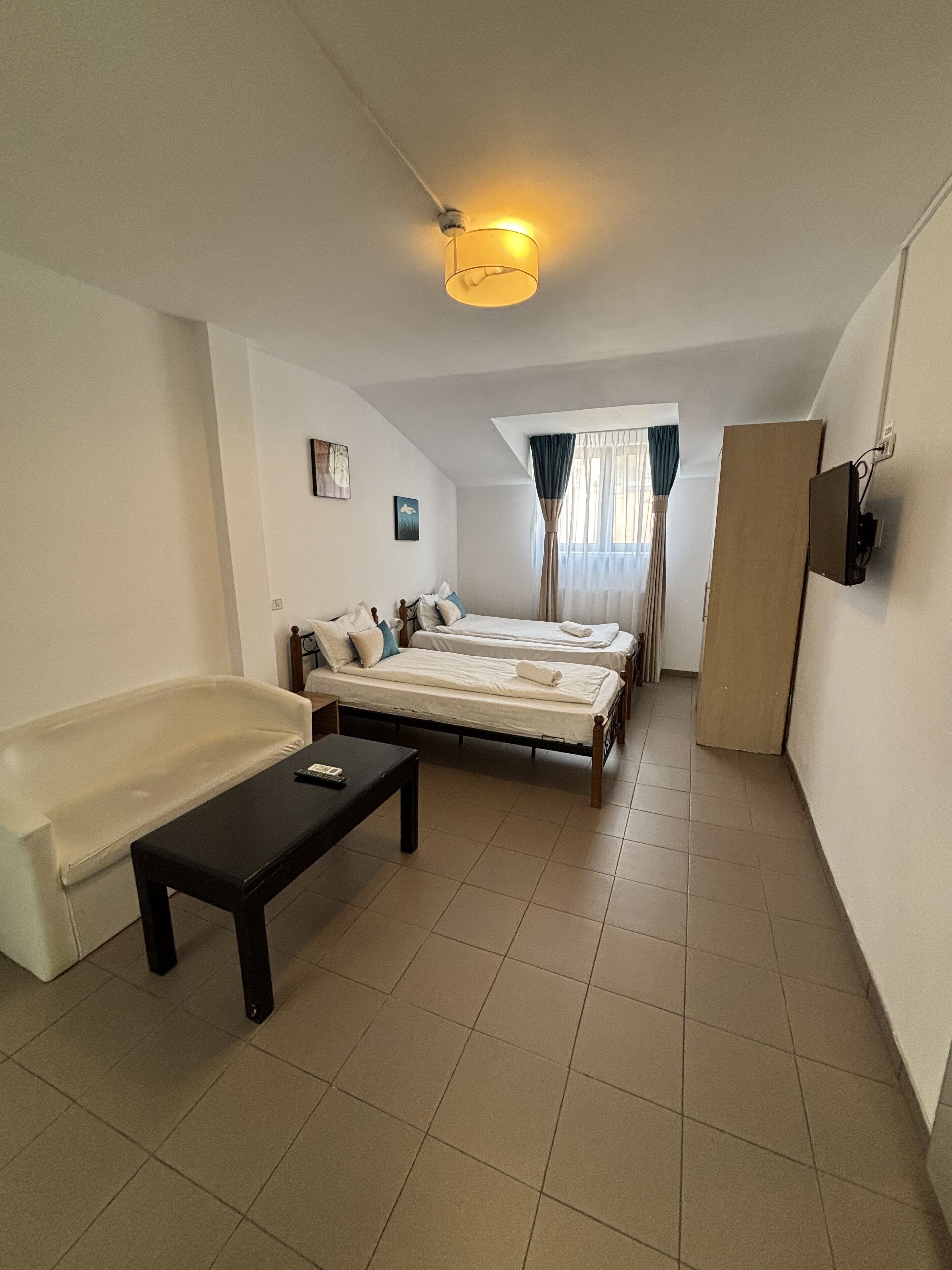 Une petite pièce bien éclairée, avec un lit simple, un canapé, une petite table et un téléviseur fixé au mur.