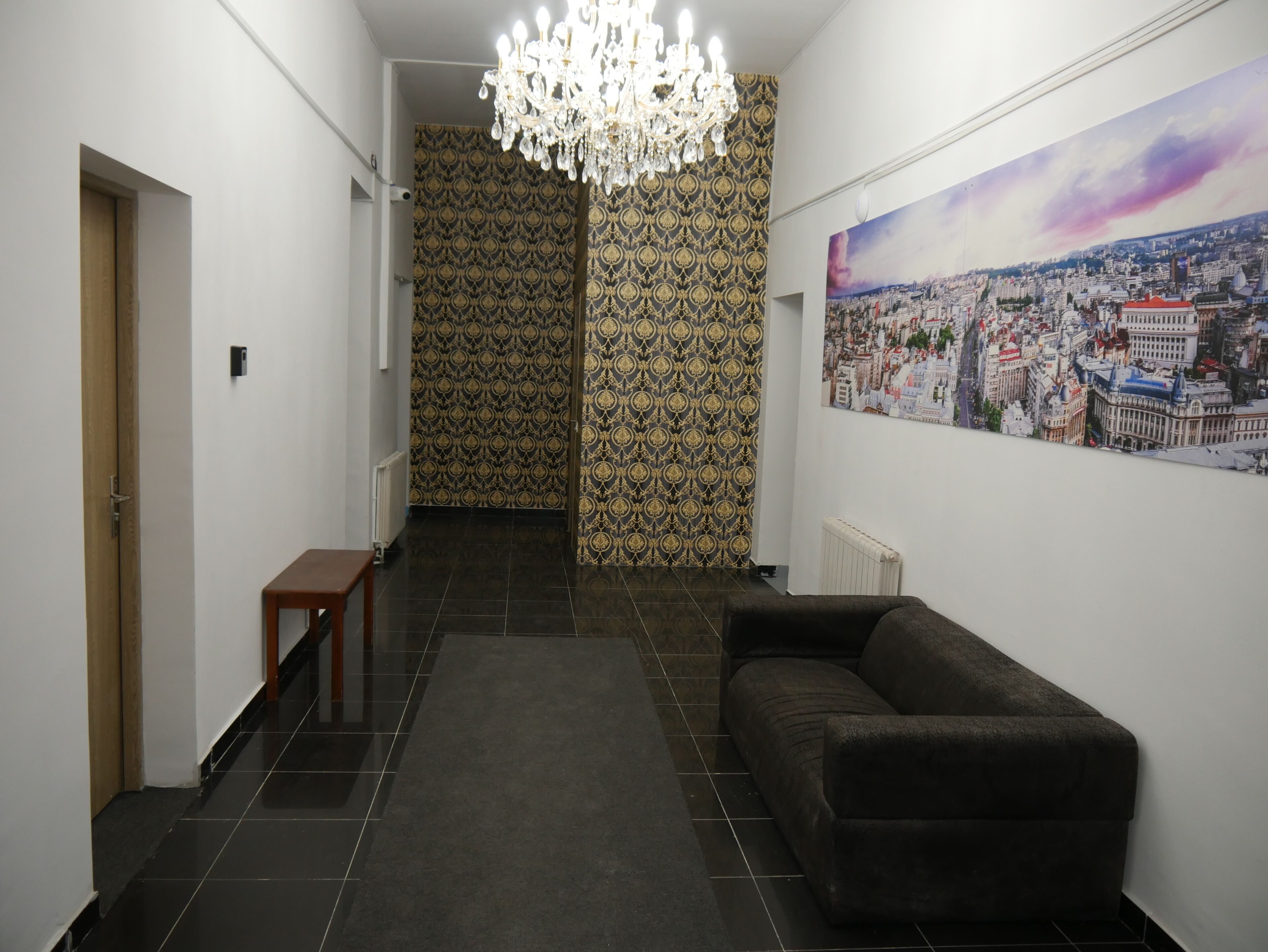 Ein Flur mit dunklen Fliesen, einem braunen Sofa, einer Holzbank, einer Chandelierleuchtre und einem Stadtbild an der Wand.