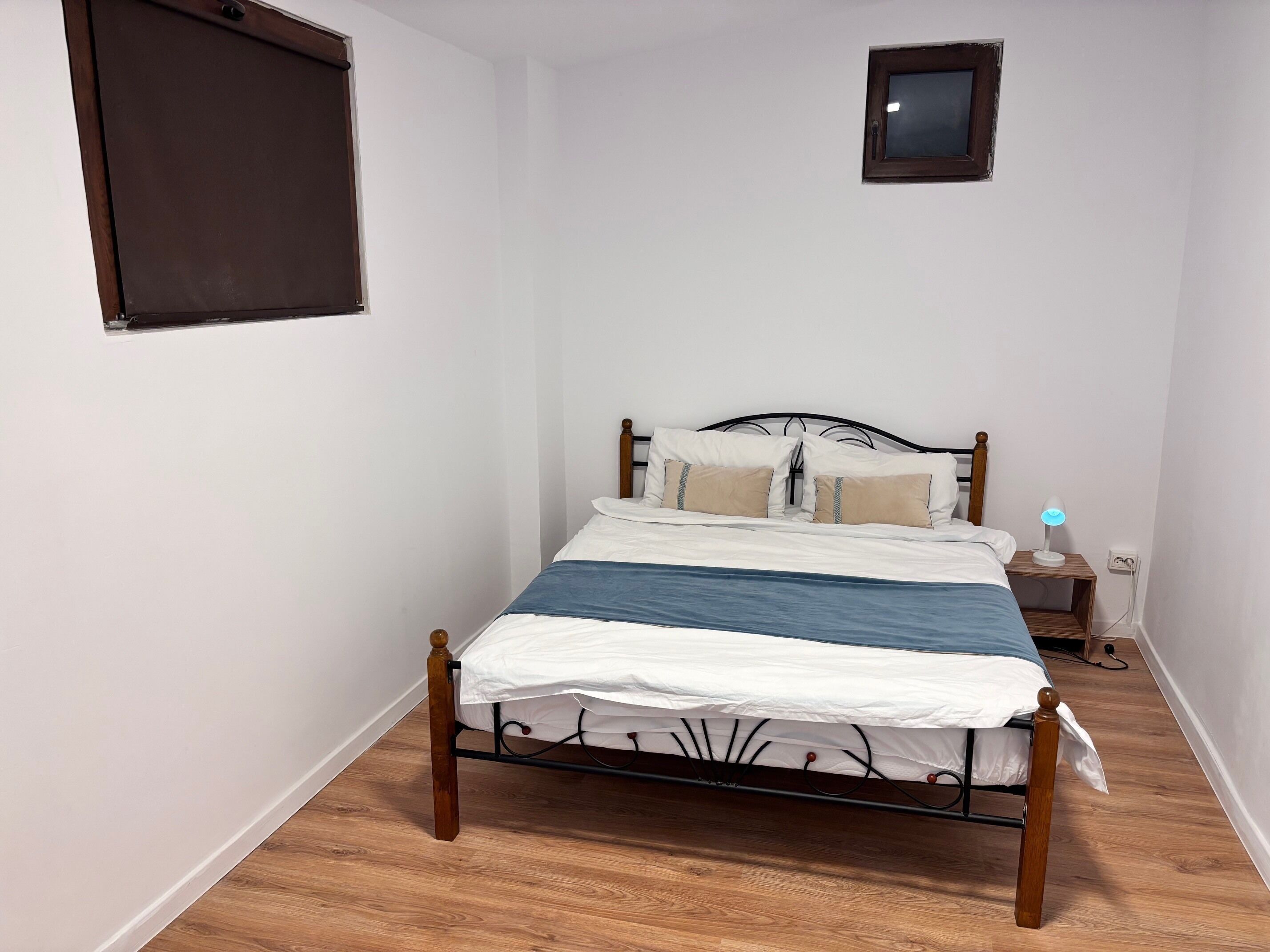 A bedroom with a bed, a bedside table with a lamp, a small wall-mounted TV, and a window with blinds.