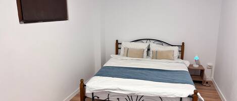 A bedroom with a bed, a bedside table with a lamp, a small wall-mounted TV, and a window with blinds.