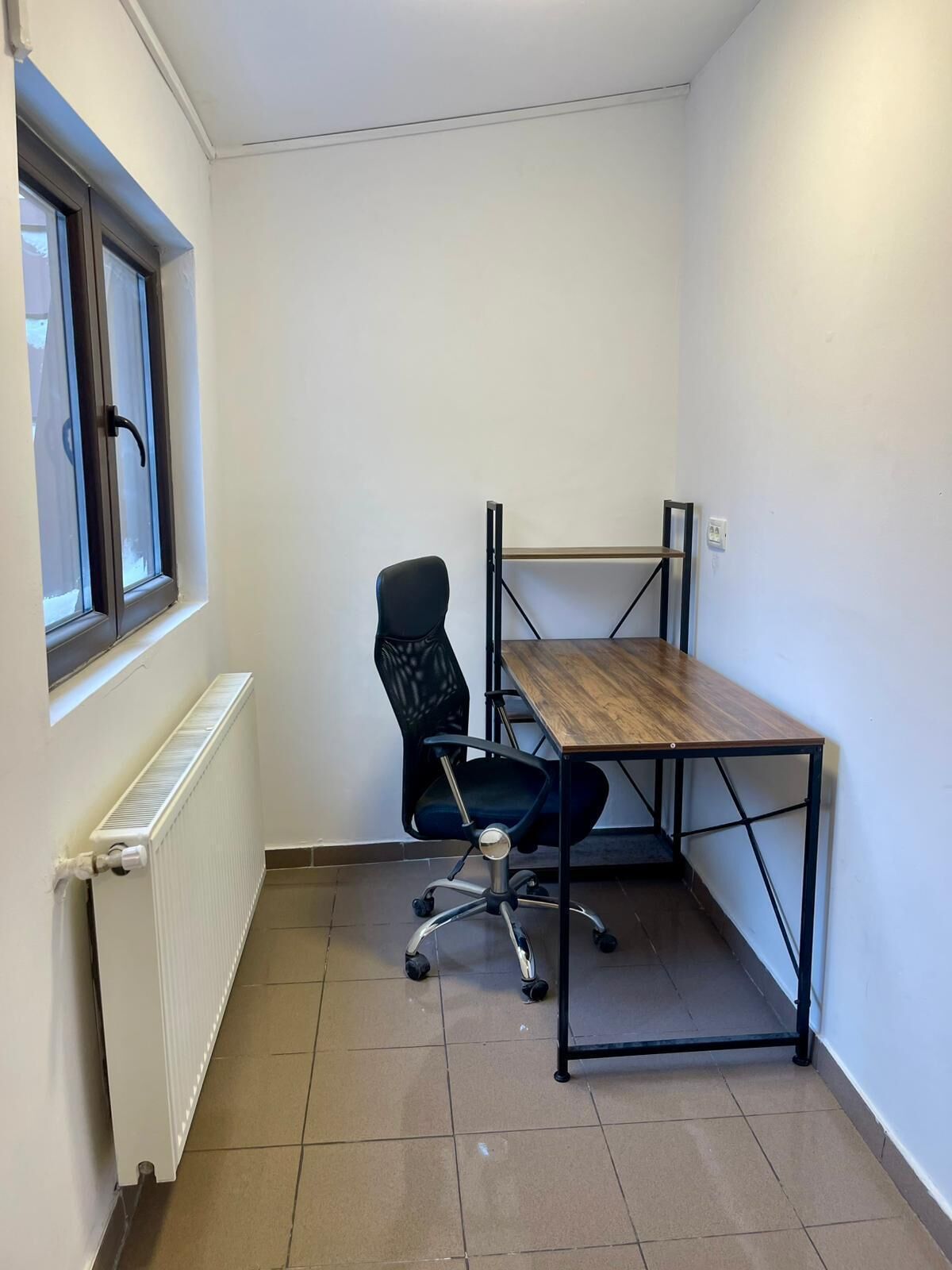 Una stanza piccola e pulita con una sedia da ufficio nera, una scrivania in legno e un termosifone.