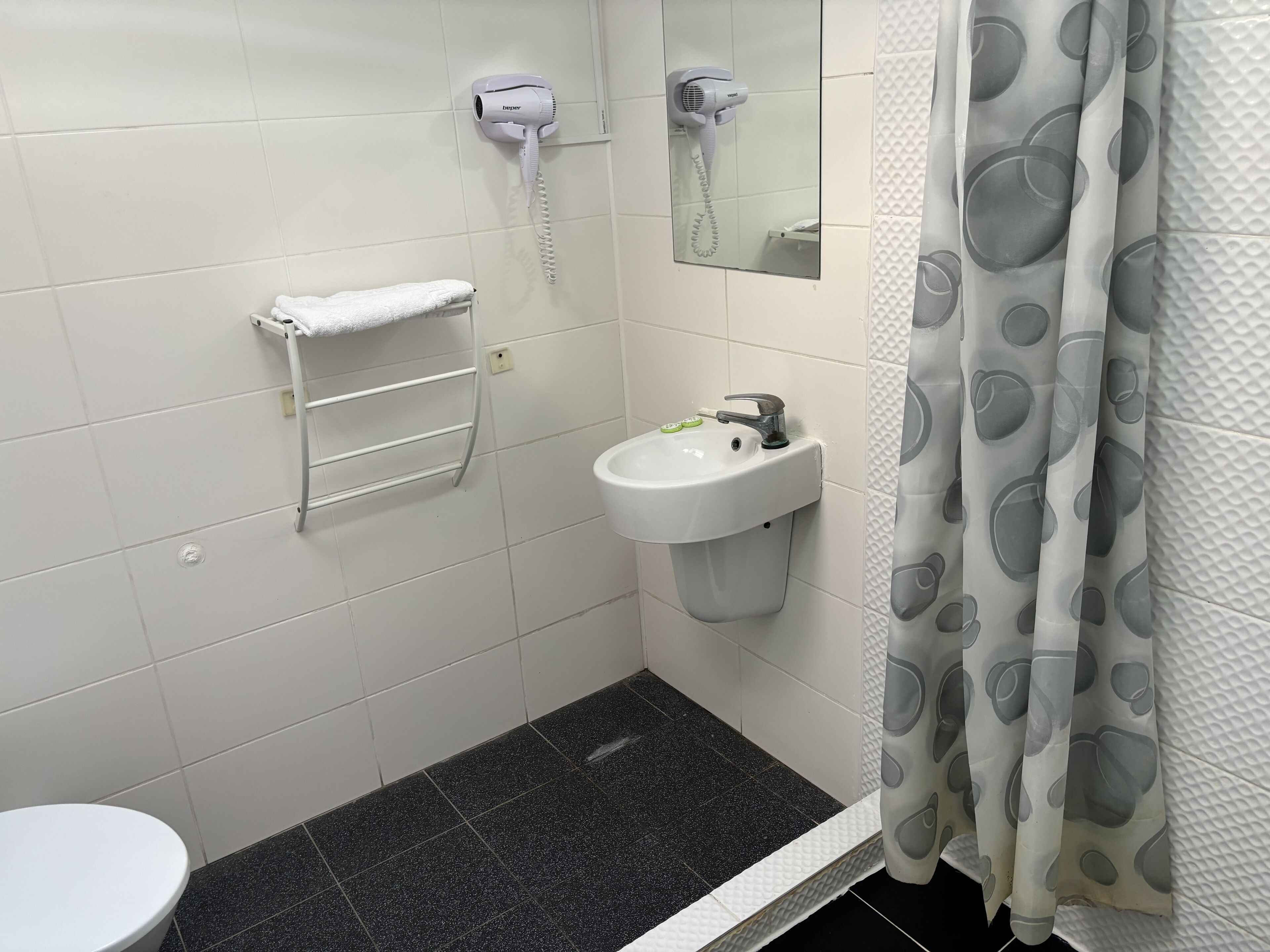 Bagno con lavandino bianco, porta asciugamani, specchio e doccia con tenda grigia.