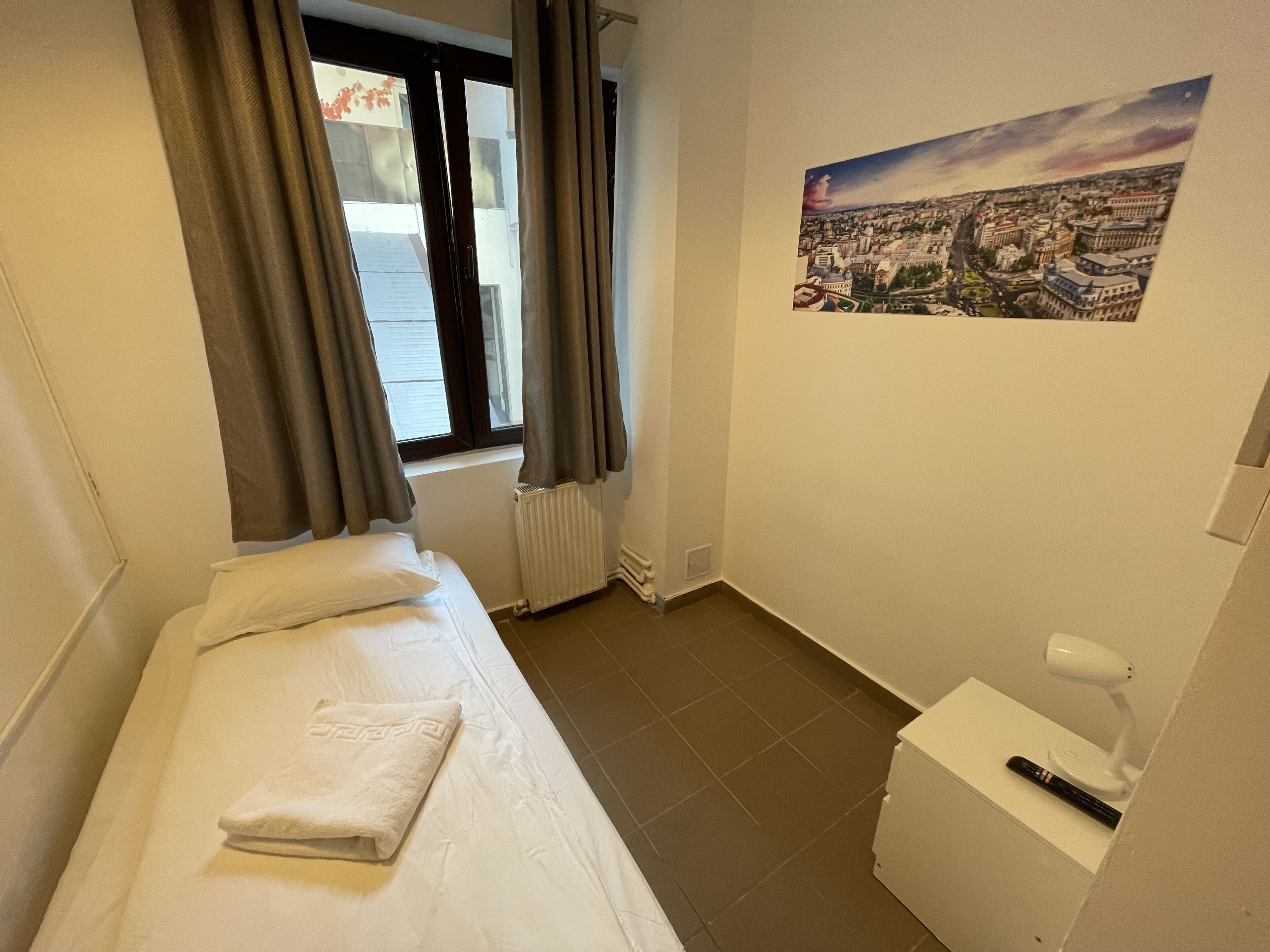 Ein kleiner, sauberer Raum mit Bett, Fenster mit Vorhängen, Heizkörper, Toilette und einem Wandbild.