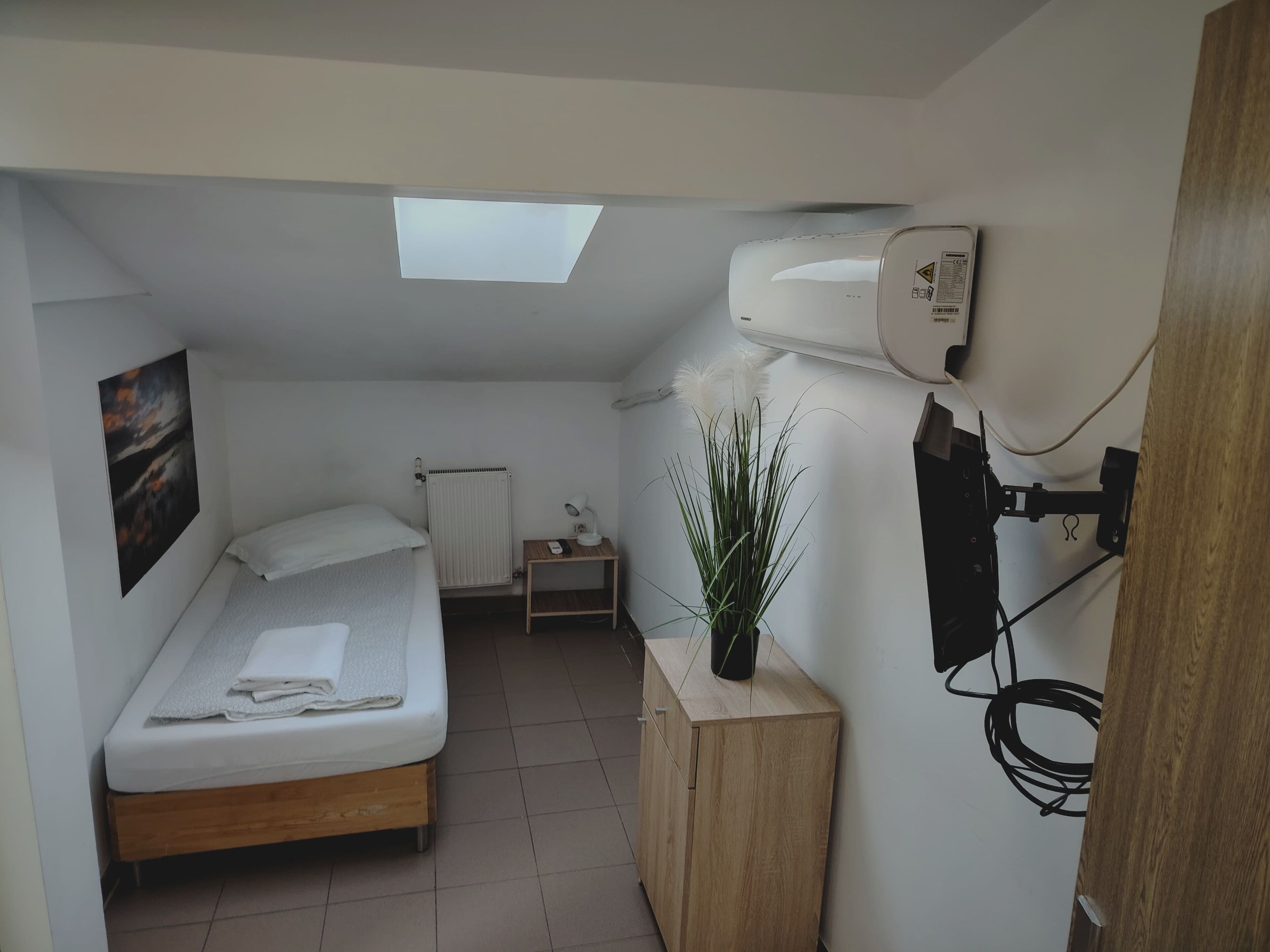 A small, modern bedroom with a bed, a radiator, a desk, a plant, and a ceiling light.