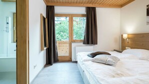 A bedroom with a wooden ceiling, a large window with curtains, a bed with pillows, and a wall-mounted lamp.