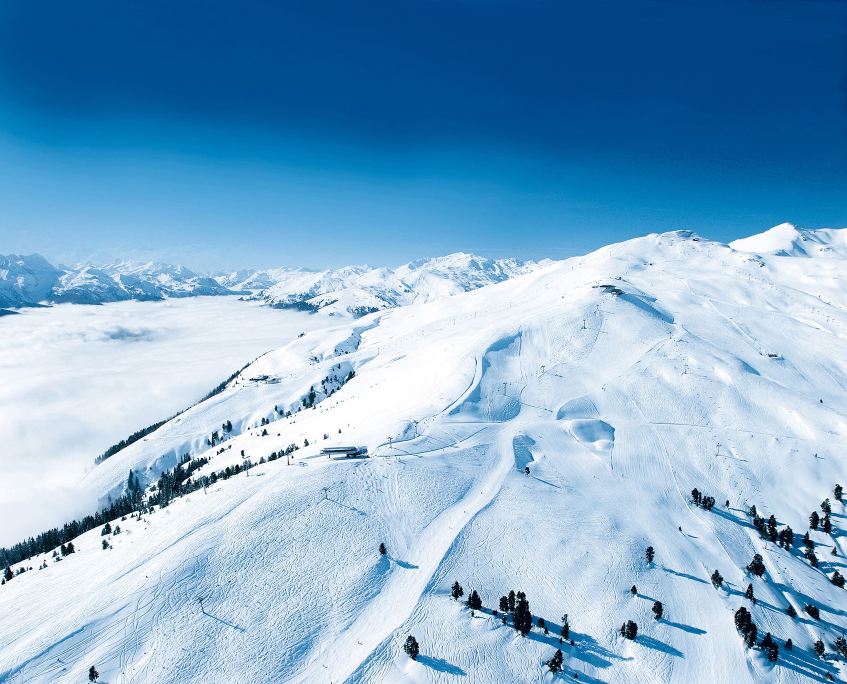 A snow-covered mountain slope with ski tracks, trees, and distant snow-capped peaks under a clear blue sky.