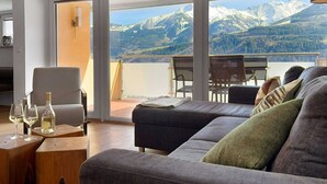 A modern living room with a large sofa, a dining table, and chairs. There are wine glasses and bottles on a side table. The room has large windows with a mountain view. - Spacious Apartment in Piesendorf near Ski Area (Piesendorf)