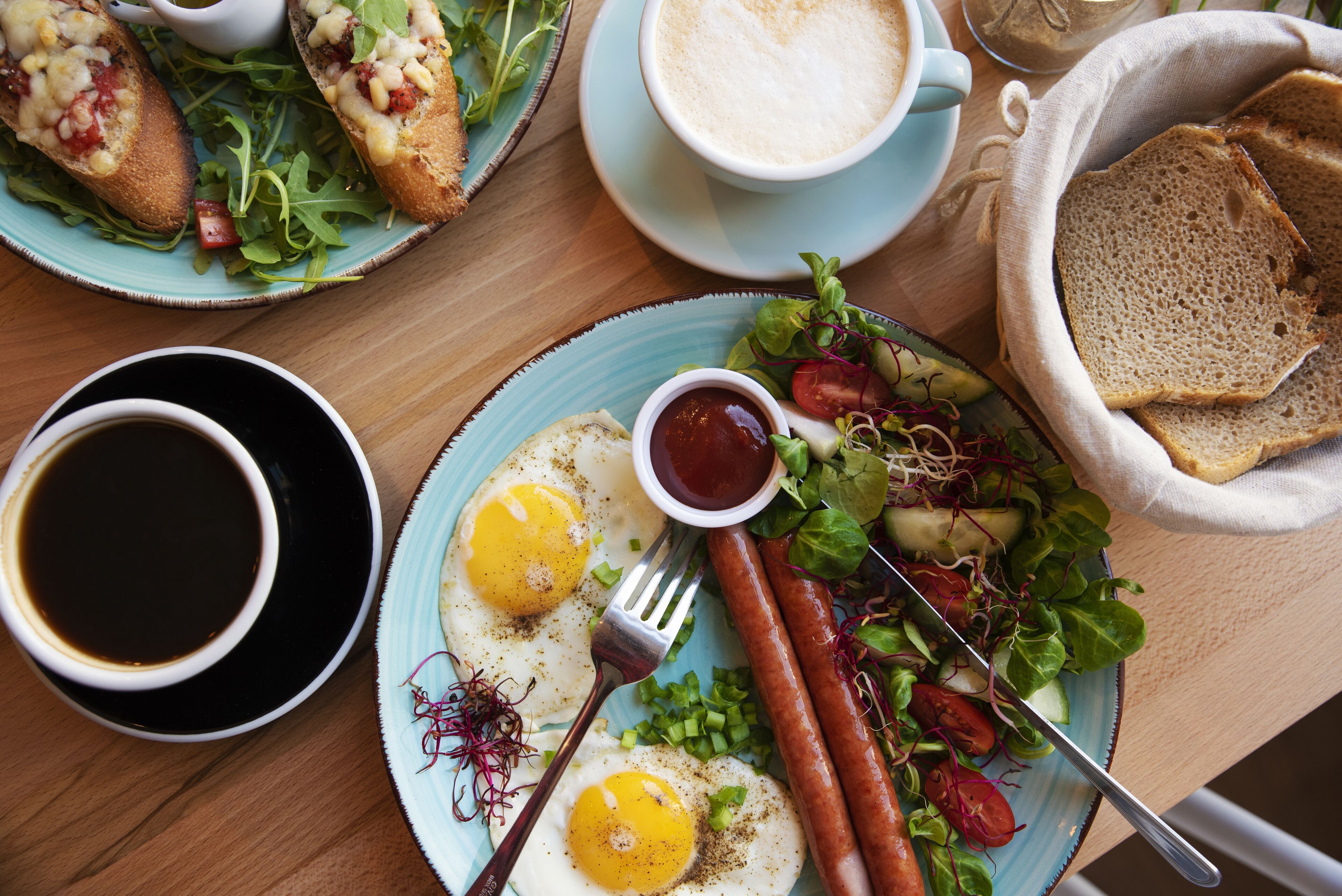 A breakfast plate with eggs, sausages, and salad, a cup of coffee, and a bowl of bread on a wooden table.