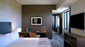 Una habitación de hotel moderna con una cama grande, un escritorio, una silla, un televisor y un cuadro en la pared.