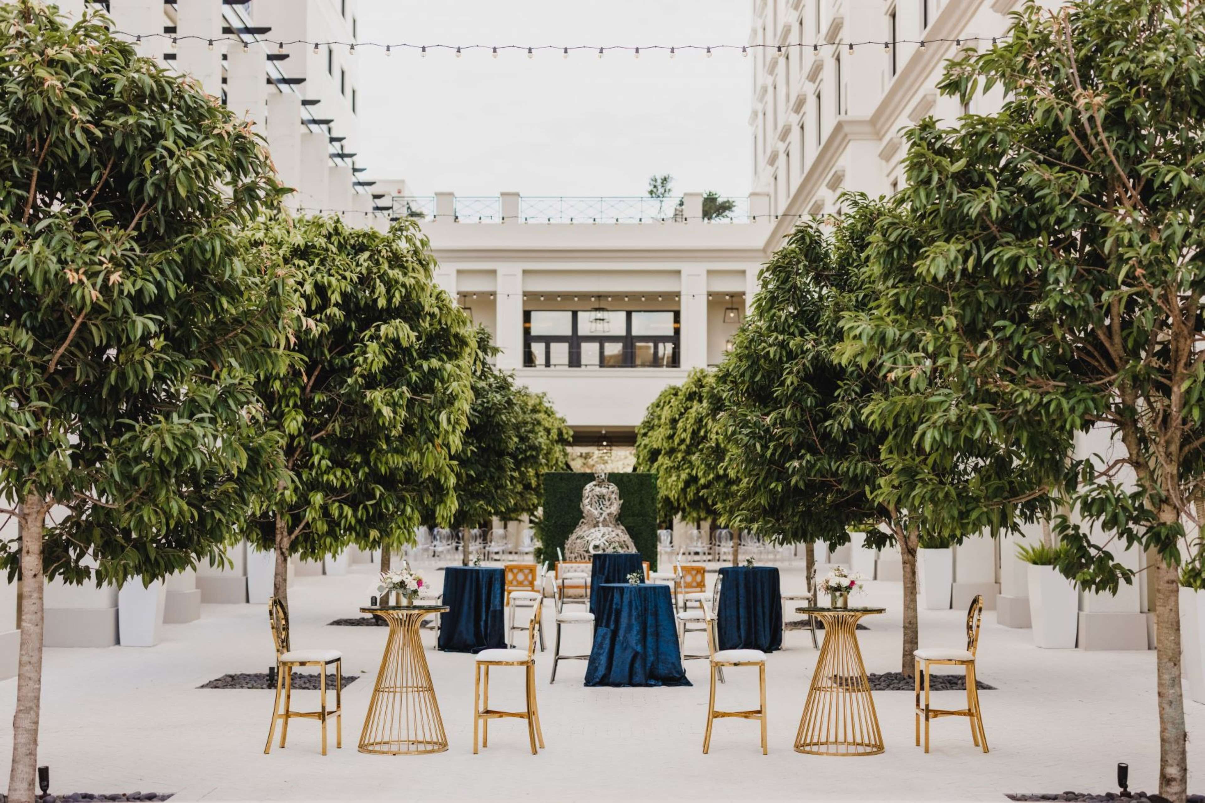 An outdoor seating area with tables and chairs, surrounded by trees and a building in the background.