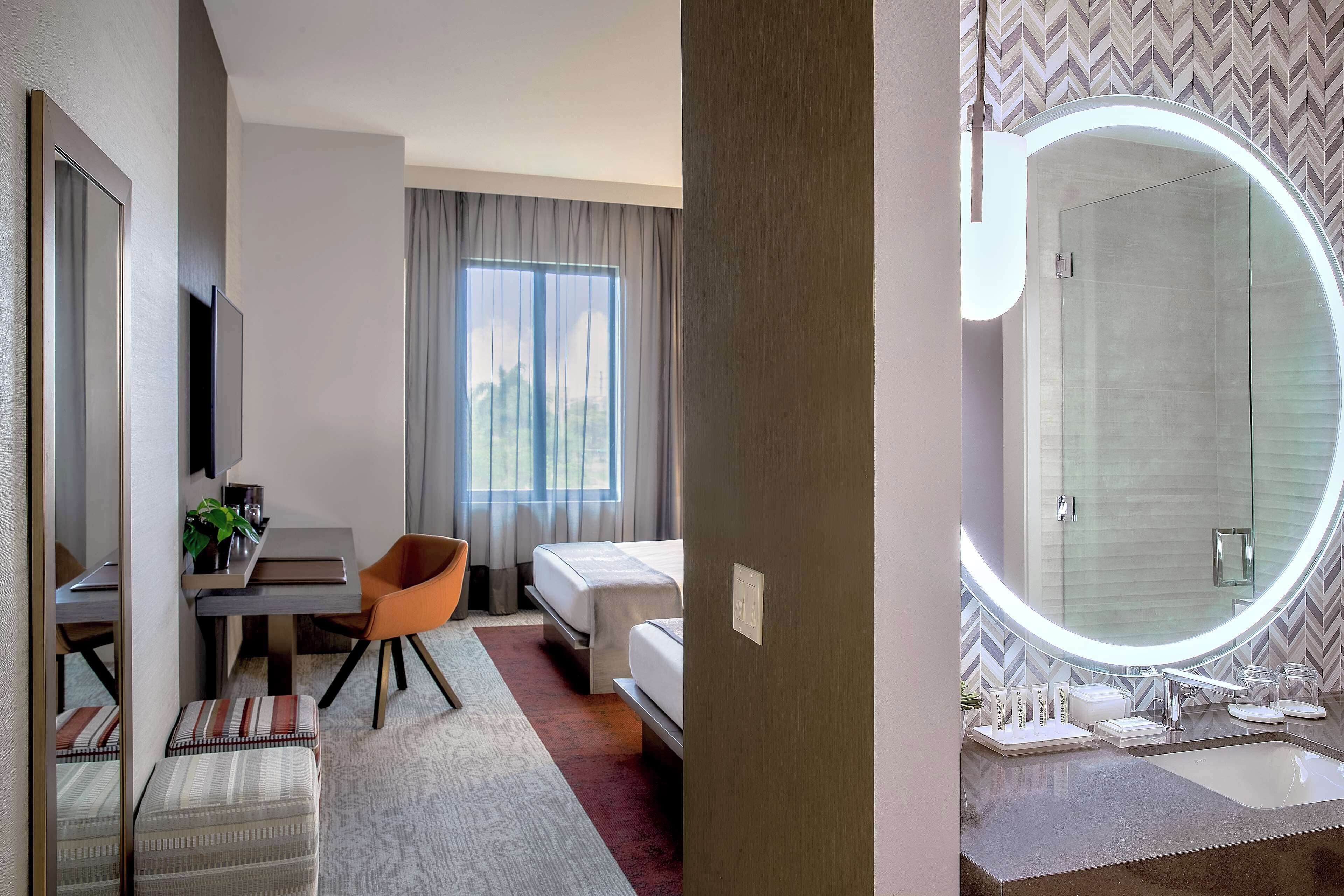 A hotel room with a bed, a desk, a chair, a small table, and a large mirror.