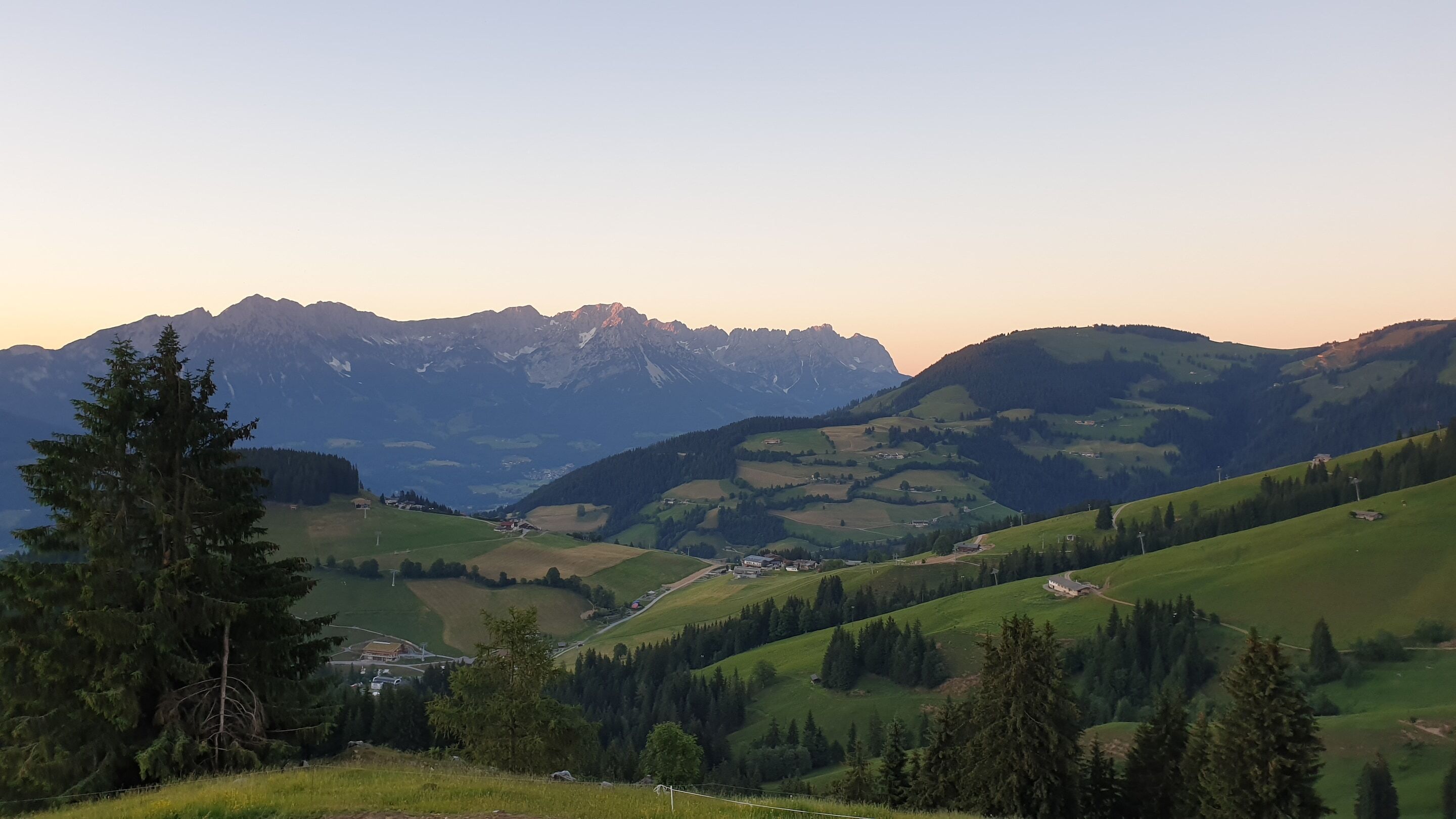 Un paysage montagneux avec une vallée, quelques bâtiments et un arbre imposant au premier plan.