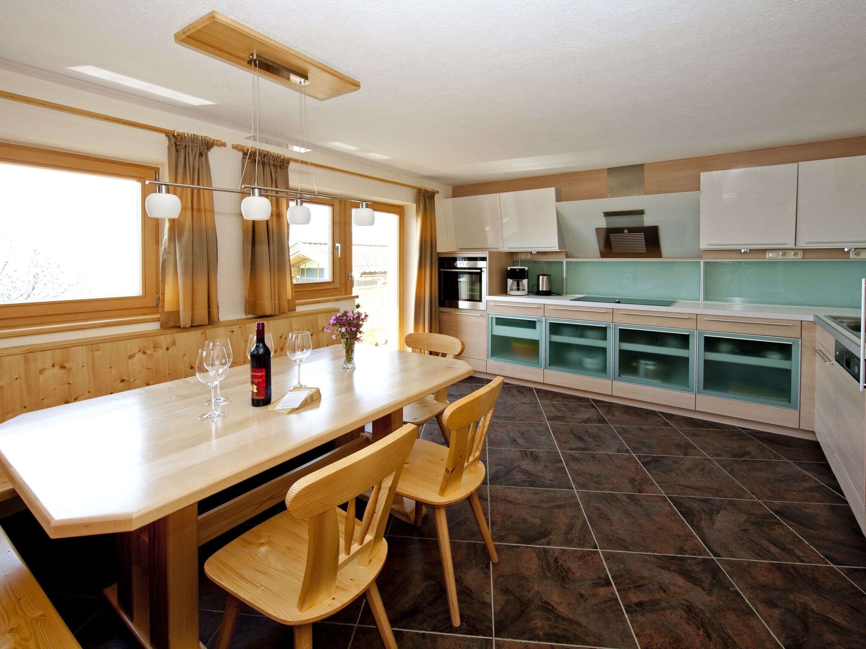 A modern kitchen with wooden cabinetry, a dining table with chairs, and a view of the outside through large windows.
