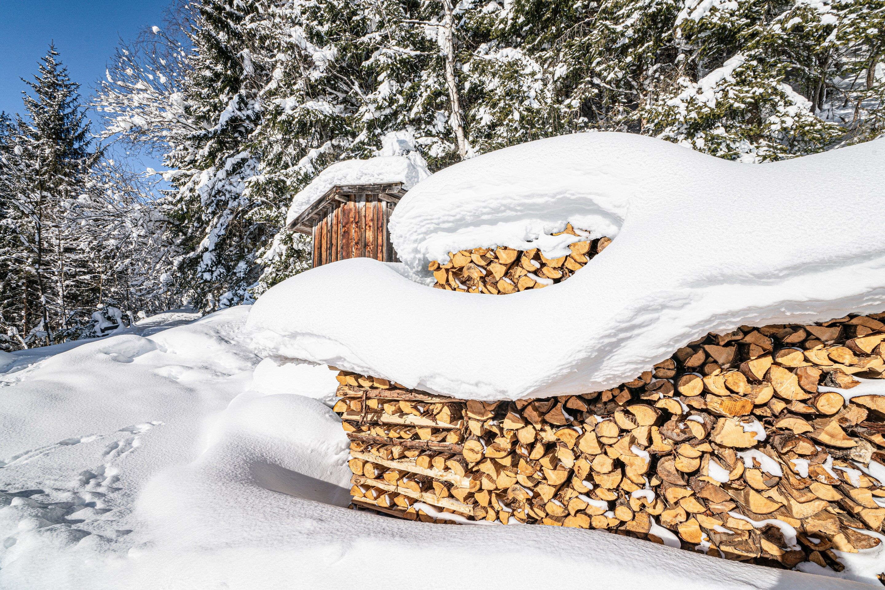 A snow-covered wooden structure with a snow-covered roof and a stack of firewood in front.