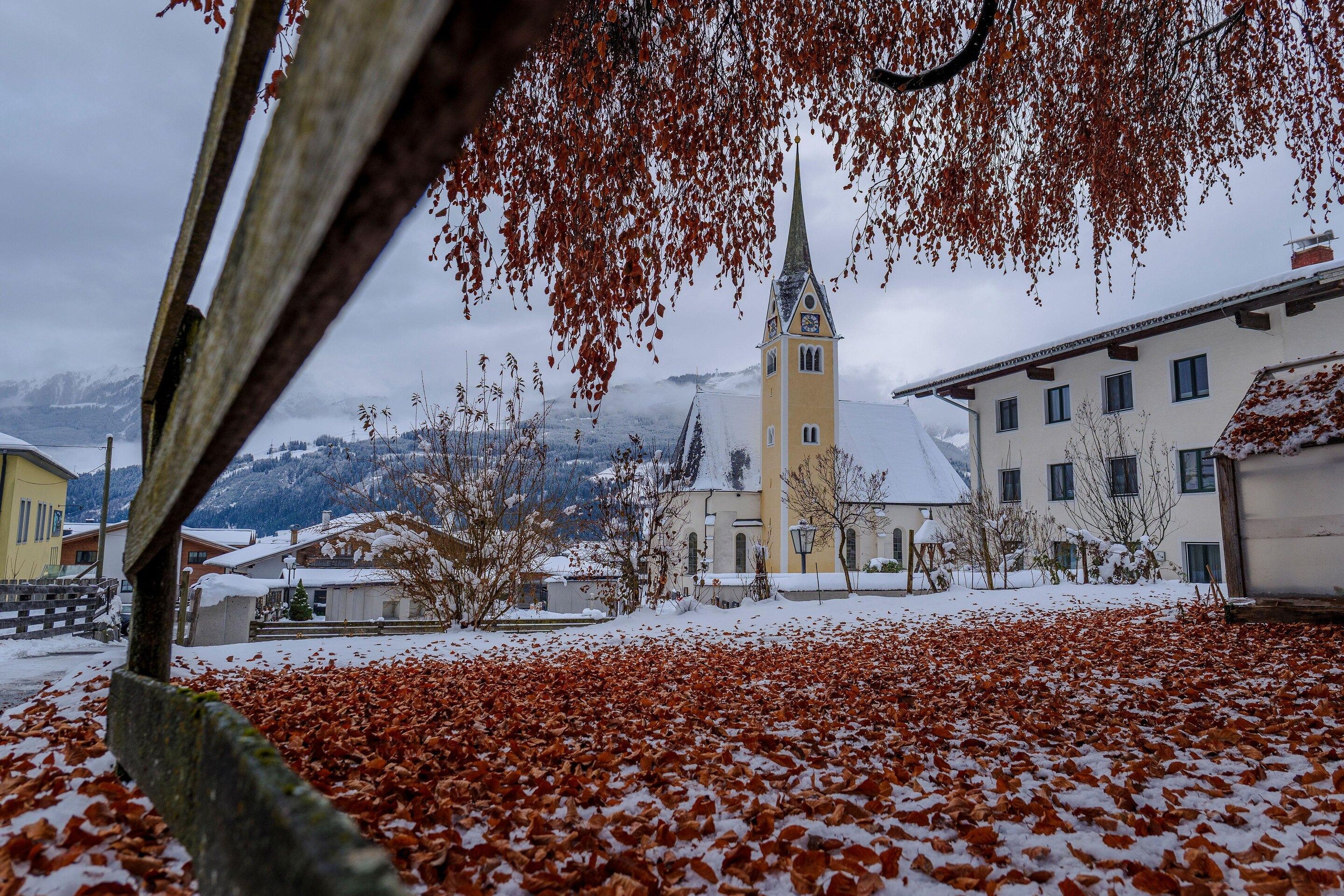 A snow-covered landscape with a church spire, buildings, and a tree with autumn leaves.