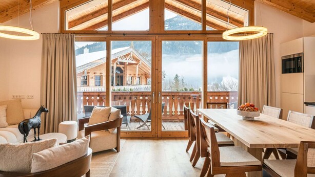 A spacious room with a high wooden ceiling, large windows, a dining area with wooden furniture, and a view of a mountain chalet. - Premium Chalet With Sauna on a Sunny Slope (Neukirchen am Grossvenediger)