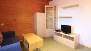 A small, cozy room with wooden walls, a blue sofa, a small table, a cabinet with glass doors, and a TV on a white stand. - Lovely Apartment in Mittersill near Kitzbühel - Kirchberg (Mittersill)