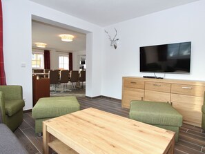 A modern living room with a wooden coffee table, green upholstered sofas, a flat-screen TV mounted on the wall, and a wooden cabinet.