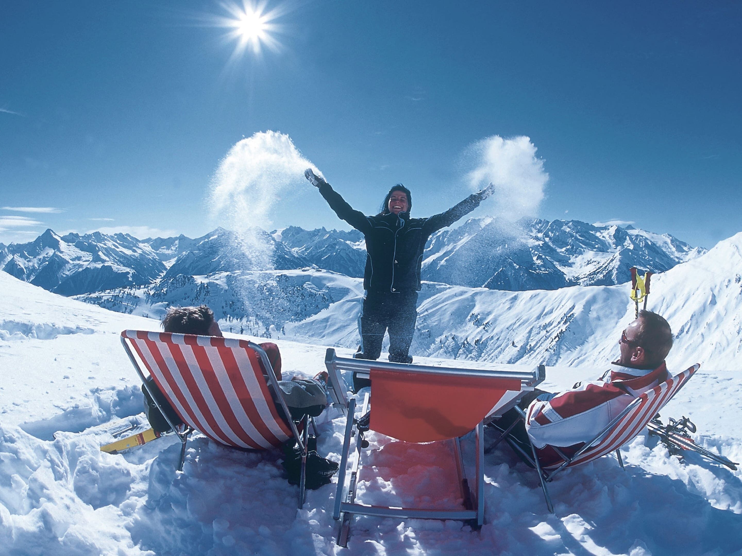 Eine Person steht in einer verschneiten Berglandschaft, die Arme in die Höhe gestreckt. Im Vordergrund befinden sich zwei rot-weiß gestreifte Stühle.