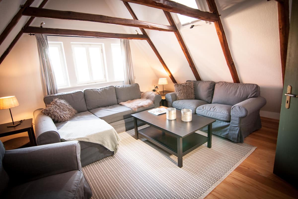 A cozy loft-style living room with exposed wooden beams, a gray sectional sofa, a small table with candles, and a view of the outdoors through a window.