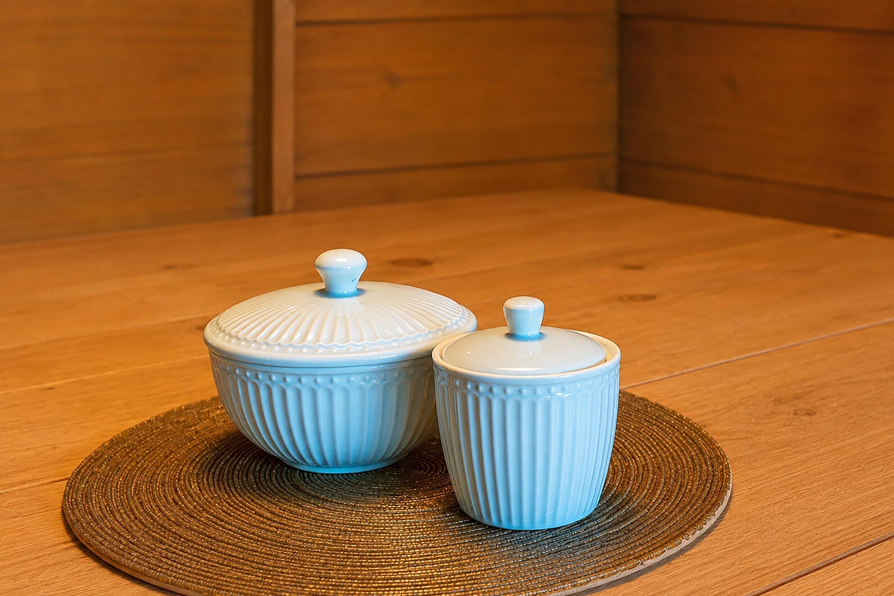 Two ceramic containers with lids on a round woven placemat on a wooden table.