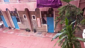 A building with a tiled ground floor and a balcony with a purple cloth. There are potted plants on the balcony and a large tree in the foreground. - Jupiter House (Kitengela)