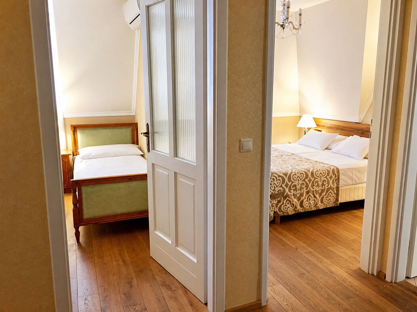 A hotel room with a wooden floor, a bed with a patterned bedspread, a bedside table with a lamp, and a window with blinds.