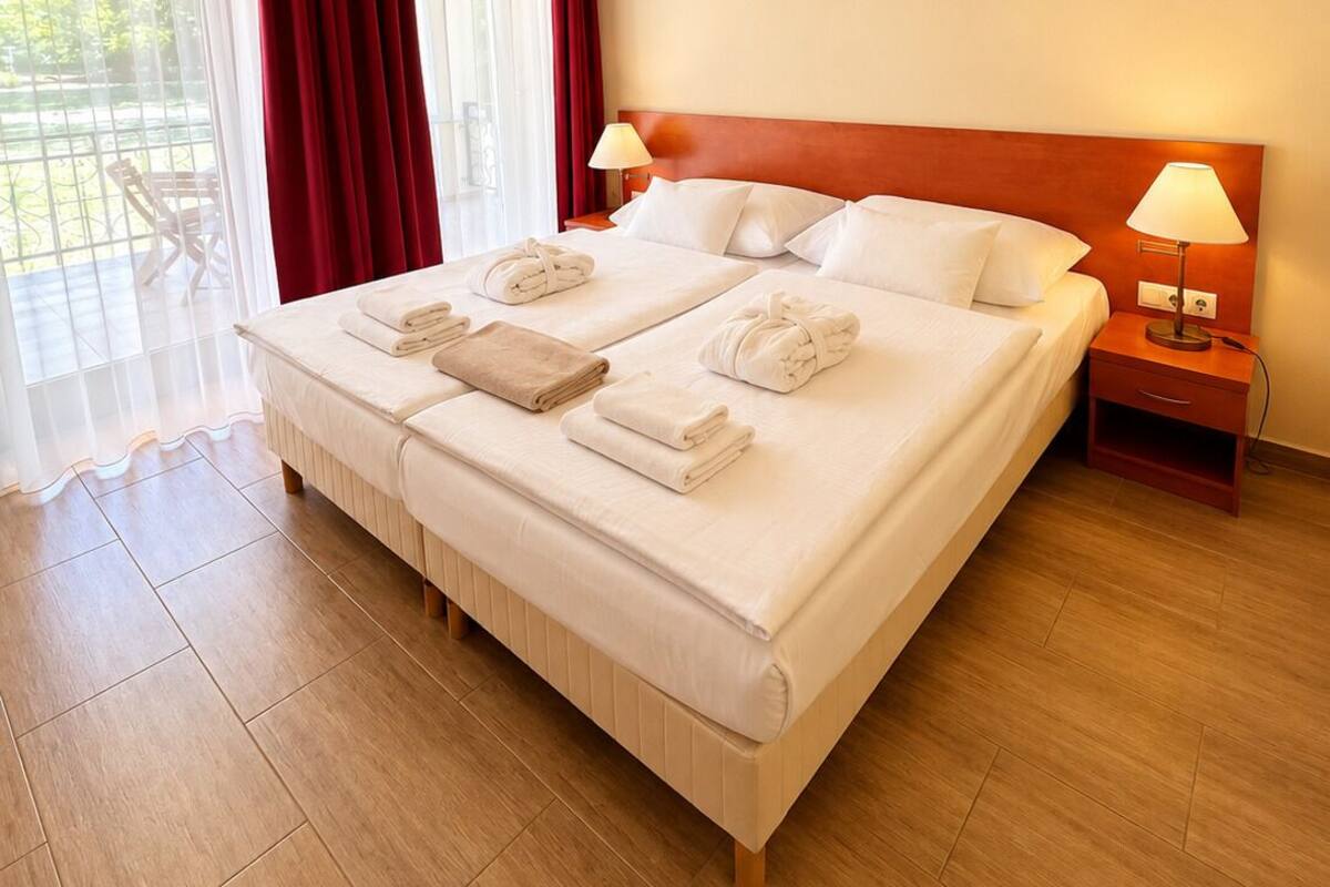 A hotel room with a large bed, two bedside tables, a window with curtains, and a view of greenery outside.