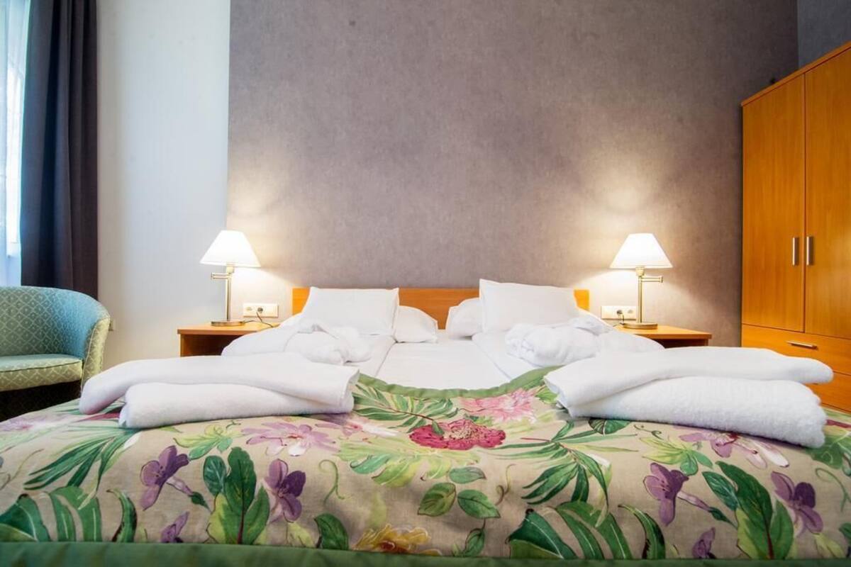 A hotel room with a bed, two bedside lamps, a wooden wardrobe, and a green armchair.